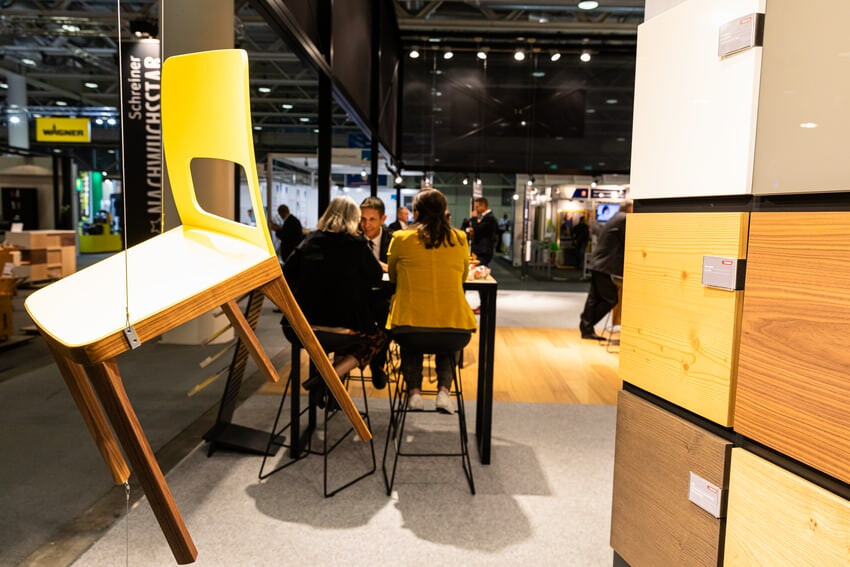 Schwebender Stuhl aus Holzplatten mit Stand-Besucher im Hintergrund.
